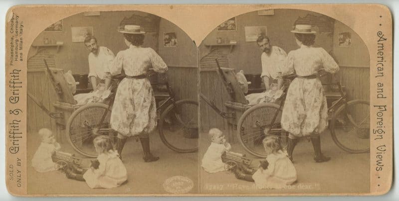Stereograph depicting a woman in breeches giving orders to a man. The woman holds a bicycle while the man is cleaning dishes. Two small children sit on the floor in the foreground.