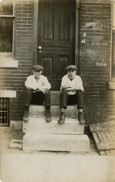 Two boys sit on a stoop. Grafitti is visible on the brick wall behind them.