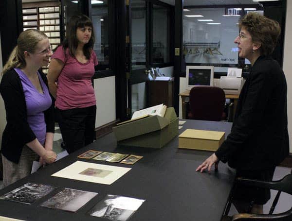 Jane Rawson, Nicole Joniec, and Sarah Weatherwax stand around a table in the Print Department. The table is laid out with a variety of collections materials that are too small to distinguish.