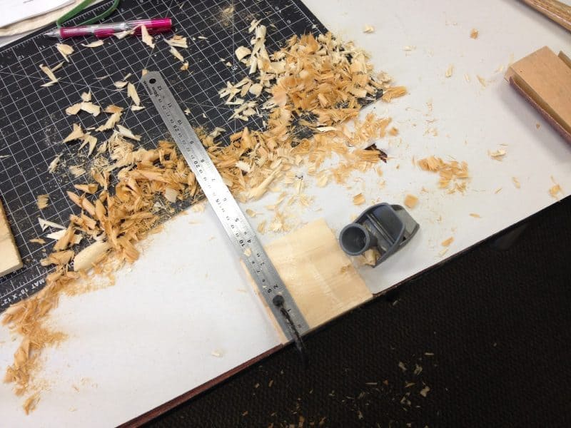 Wood shavings cover a work space. A ruler measures a small block of wood.