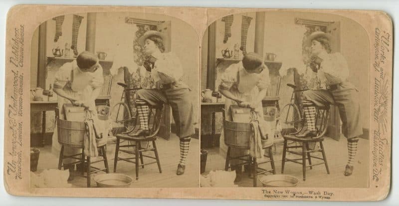 Stereograph depicting a woman in breeches watching a man do laundry. A bicycle is placed in the background. 