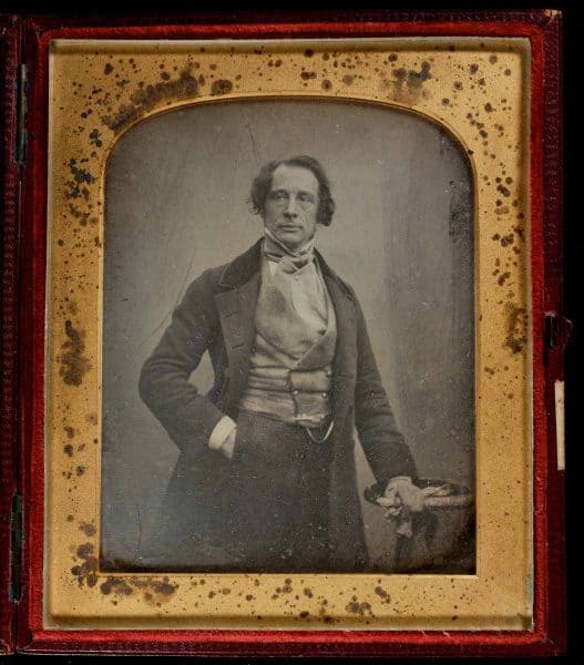 Cased photograph. Three quarter length pose of Charles Dickens standing with his right hand in his pocket and his left hand, holding his gloves, resting on an urn.