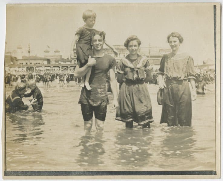 Photograph. Group of three adults and one child in knee-deep water, presumably at the beach.