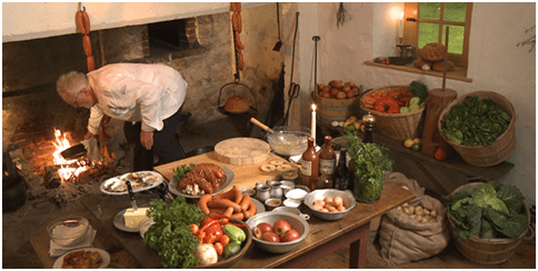 Presumably the set of “A Taste of History.” A table is laid out with a variety of foods in front of an open hearth. A man, presumably chef Walter Staib, is bent over the hearth at work.