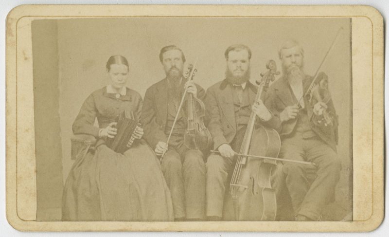 Picture shows one woman and three men, seated next to each other, and holding instruments. The woman holds an accordion in her lap and she looks slightly down. To her left is a man, his eyes closed, who holds a viola perpendicular to his lap with one hand and a bow in his other. To his left is a man resting a cello between his legs. He holds a bow across the base of the cello with his right hand. To his left is the last man, his eyes closed, who holds a violin by his left shoulder and a raised bow in in his right hand. The woman, as well as the man who holds a cello, wear glasses. The woman wears a dark dash colored corseted dress with long sleeves and a long skirt. The men, who look toward the viewer, are bearded and wear dark dash colored suits.  [End of description]