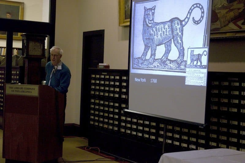 Keynote speaker Peter Benes at the podium. A projector screen to the side depicts a print of a cat.