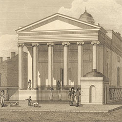Six-columned classical style building surrounded by fence. Male pedestrians and two dogs gather outside the building.