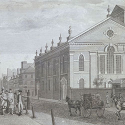White man gestures with his arm while leading group of Indigenous people along sidewalk. Horse-drawn wagons and other pedestrians pass by large buildings including a church