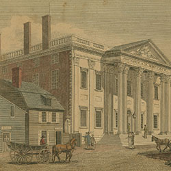 Two-storied Classical style building with columned front entrance stands near smaller buildings along cobblestoned street. Horse-drawn vehicles and pedestrians pass by buildings