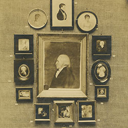 Framed waist-length profile portrait hanging on wall surrounded by smaller framed portraits