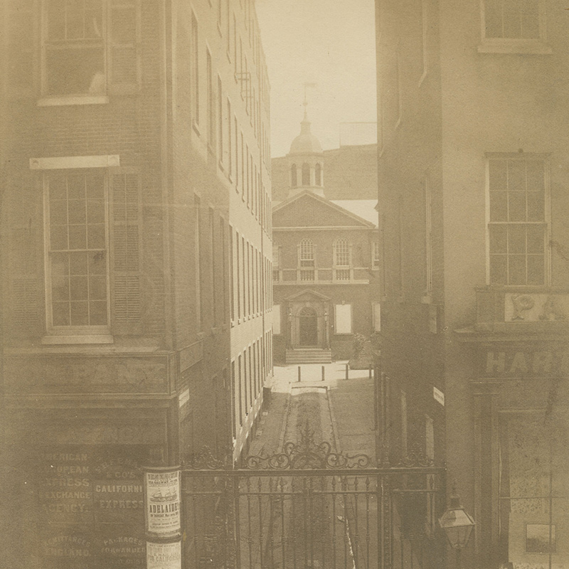 View through an alley of brick building with a steeple of Carpenters’ Hall. Portions of storefronts are visible.