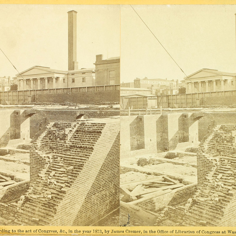 Photographic view of partial brick and stone foundations within a large opening in the ground. Buildings line the upper ground. 