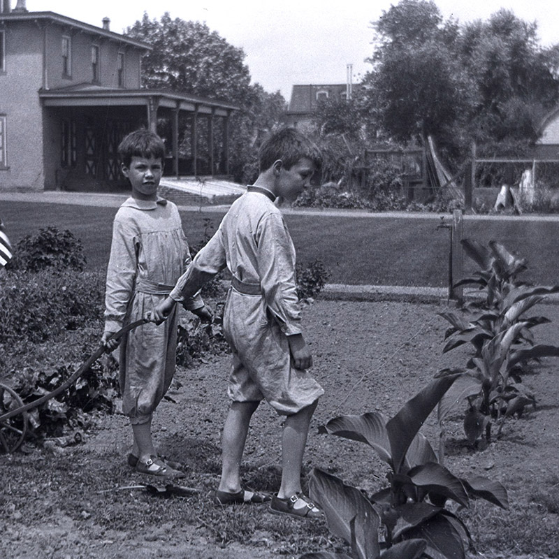Portrait of two white boys attired in jumpers and pulling a full wagon in a vegetable garden in front of a two-story house with covered back porch.