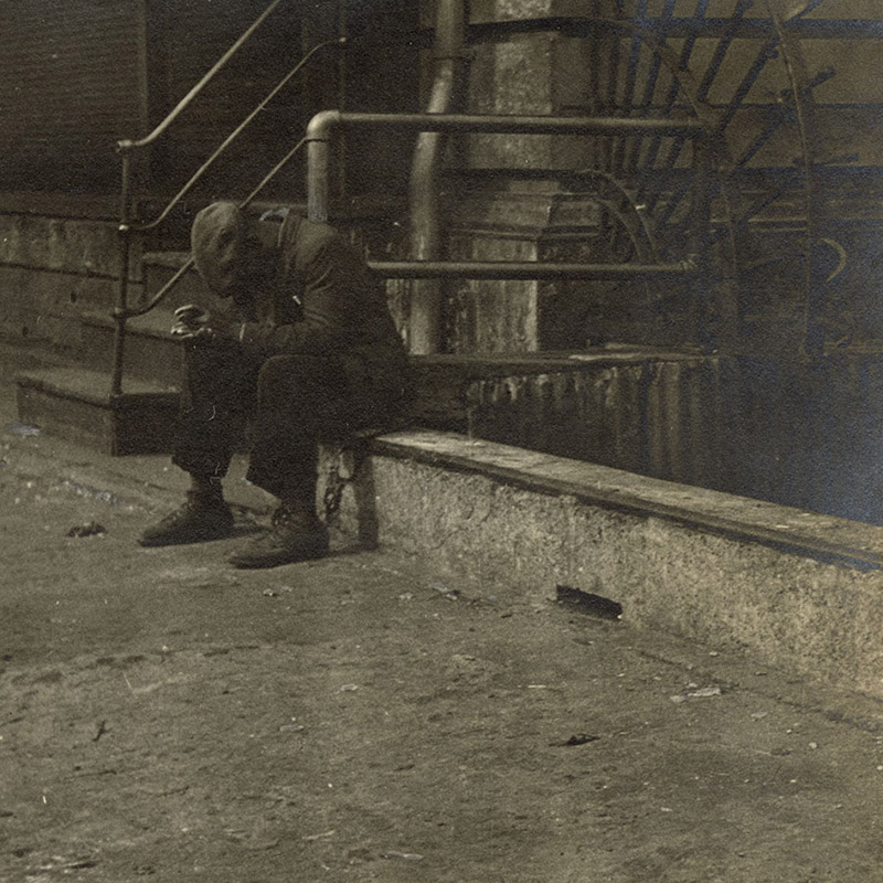 View of a Black man, his head down, and seated on embankment next to a stairwell to a building adorned with ornamental ironwork.