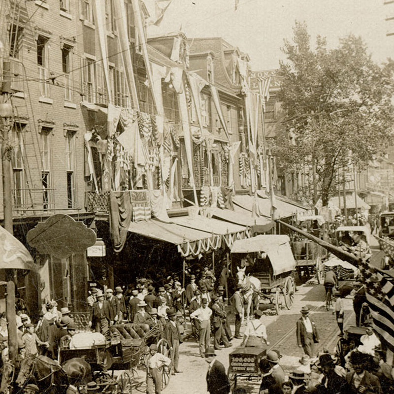 Street view depicting storefronts covered in American banners and flags alongside Race Street congested with people and horse-drawn wagons.