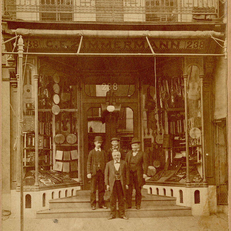 Photograph showing a group of white men in front of a music store with large display windows. String, wind, and percussion instruments hang in the windows.