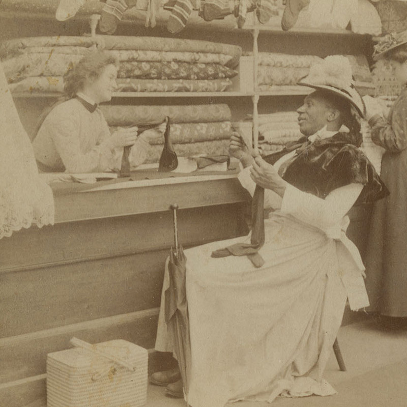 Racist caricature of seated, Black man dressed as a woman and  buying stockings from a  white woman clerk behind a counter.