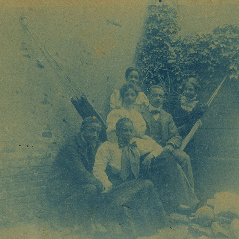 Blue color photograph of Black men and women seated on and around a hammock in a quaint city backyard.