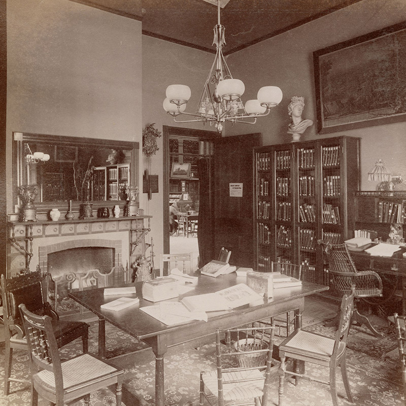 Interior view showing chairs around a table, bookcases, a wall mirror and clock, and large framed print.