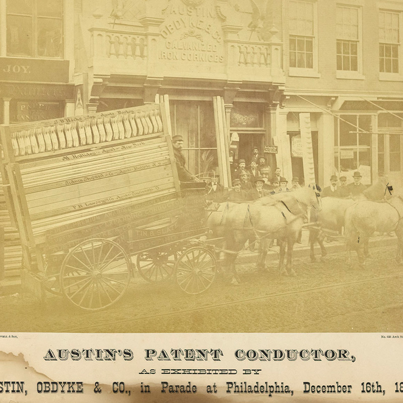 Photograph with stained and mildewed mat depicting a horse-drawn wagon display of s-shaped metal rain spouts and rectangular roof slats.