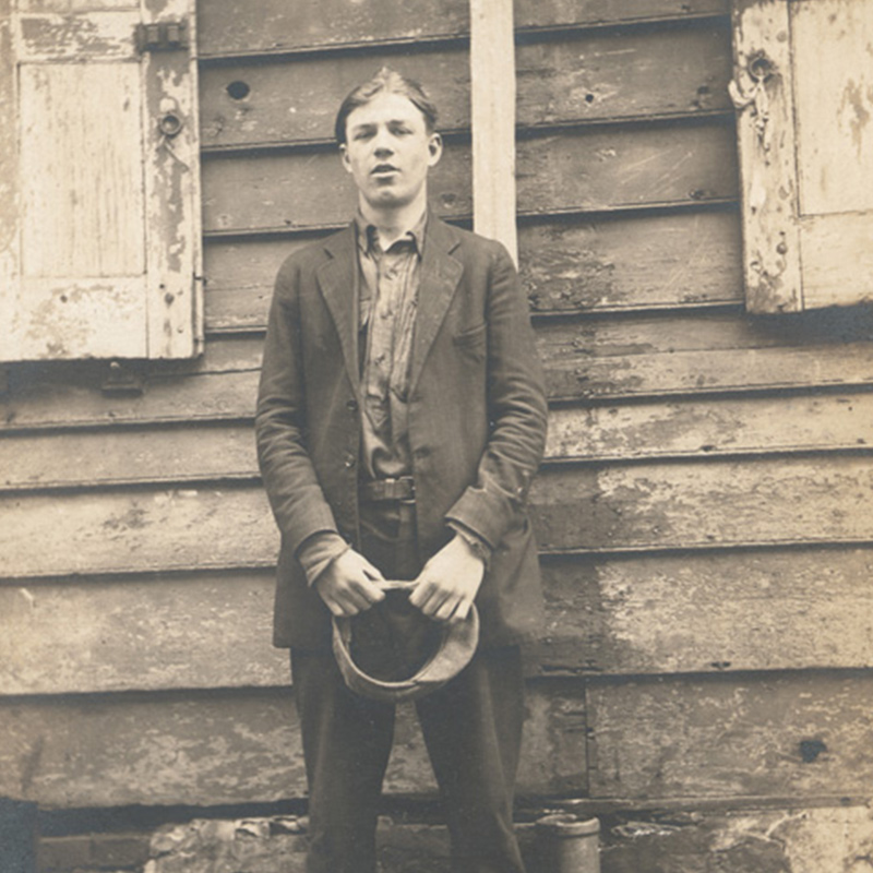 Full-length portrait of young white man, standing, holding a hat and attired in a shirt, jacket, and pants. 
