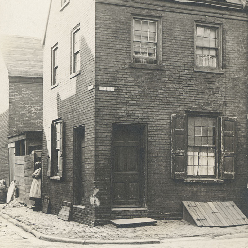 View of Black woman standing next to a two and a half story brick residence with outside door to a basement cellar.