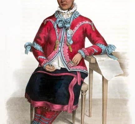 Native American woman wearing a hat, jacket and skirt over a blouse and leggings, her elbow resting on a table.