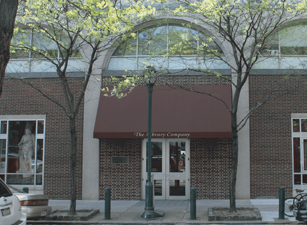 Visit button: Front doors, awning, and windows of the Library Company brick building on tree-lined Locust Street.