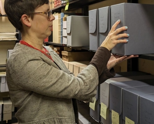 Photograph of a white woman with short brown hair and glasses putting a box of archival items back on a shelf