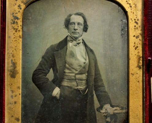 Charles Dickens with right hand in pocket and left hand resting on a small table.