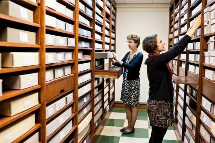 Graphic Arts Department Two white women Library Company curatorial staff members in Graphic Arts storage area reach and pull flat boxes from shelves.