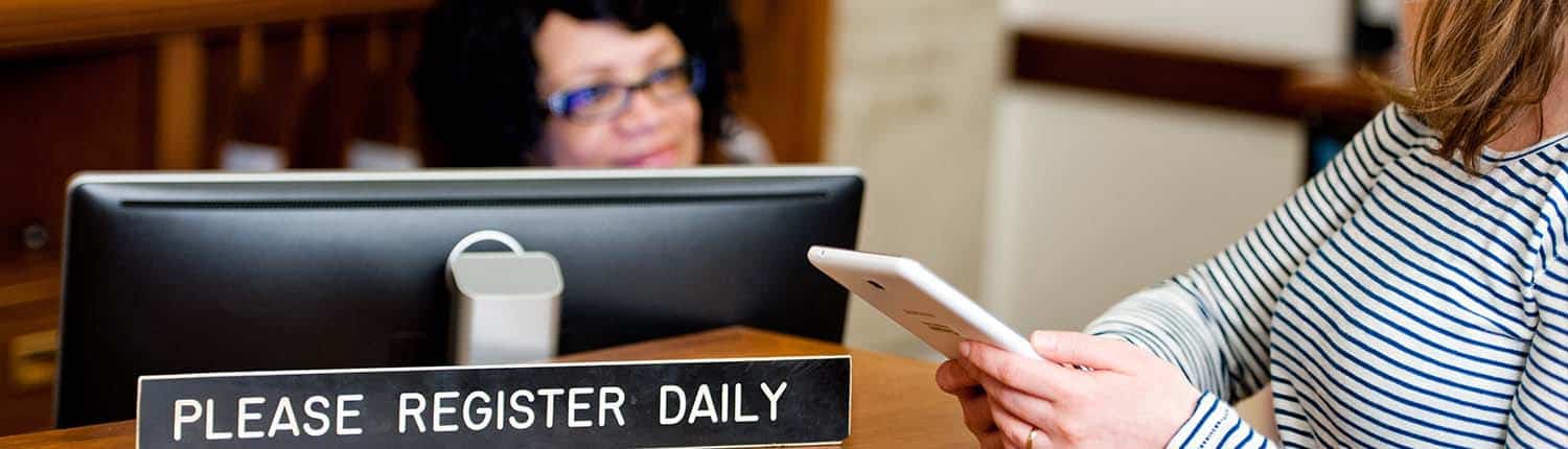 A Black woman Library Company staff member in glasses looks at white woman holding smartphone near placard please register daily in Library lobby.