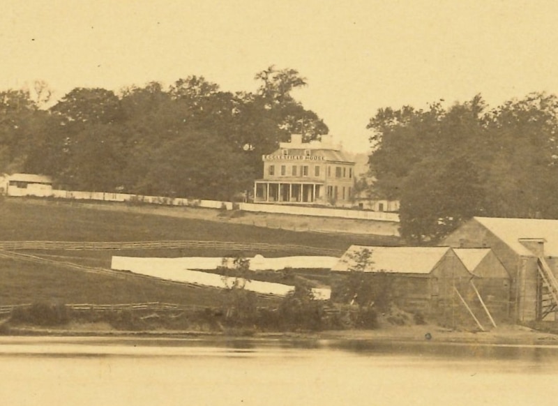 Detail of photograph showing large house off in the distance surrounded by trees, fence, outbuildings, and fields. The house has a large sign at the roofline reading Eaglesfield House.